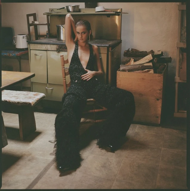 A stylish woman with dark hair sits in a chair wearing a shimmering black jumpsuit in a vintage-style room with rustic decor.