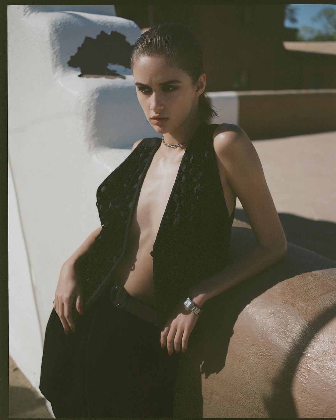A young woman poses outdoors in a black vest and dark pants, leaning against a light-brown adobe wall under natural lighting conditions.