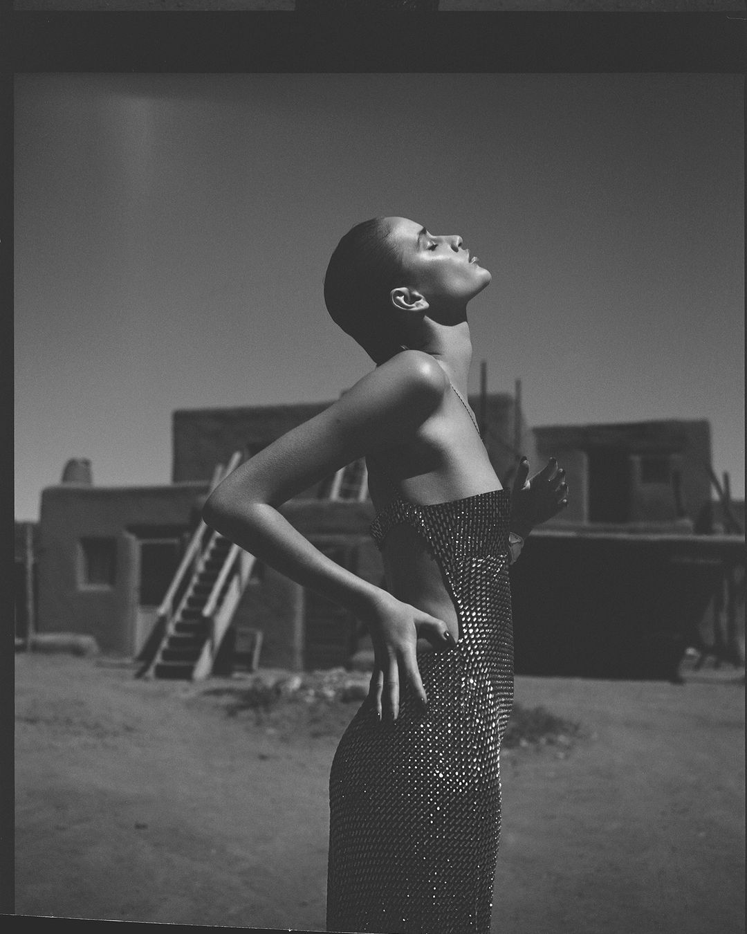 Black and white portrait of a woman with a shaved head in a sequined dress, looking up with arms at her side, against an adobe building backdrop.
