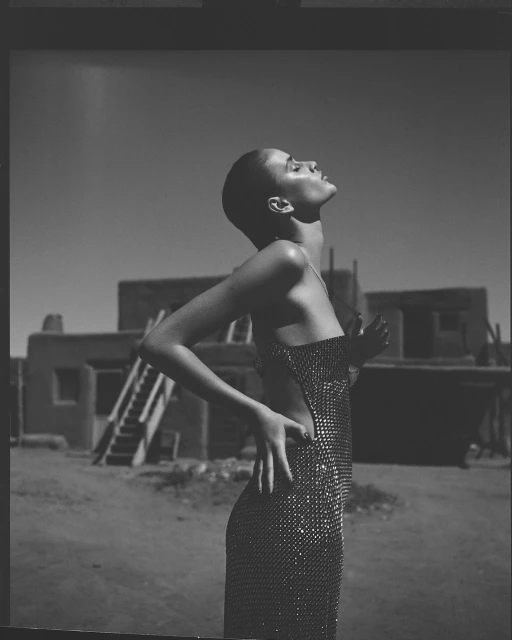 Black and white portrait of a woman with a shaved head in a sequined dress, looking up with arms at her side, against an adobe building backdrop.