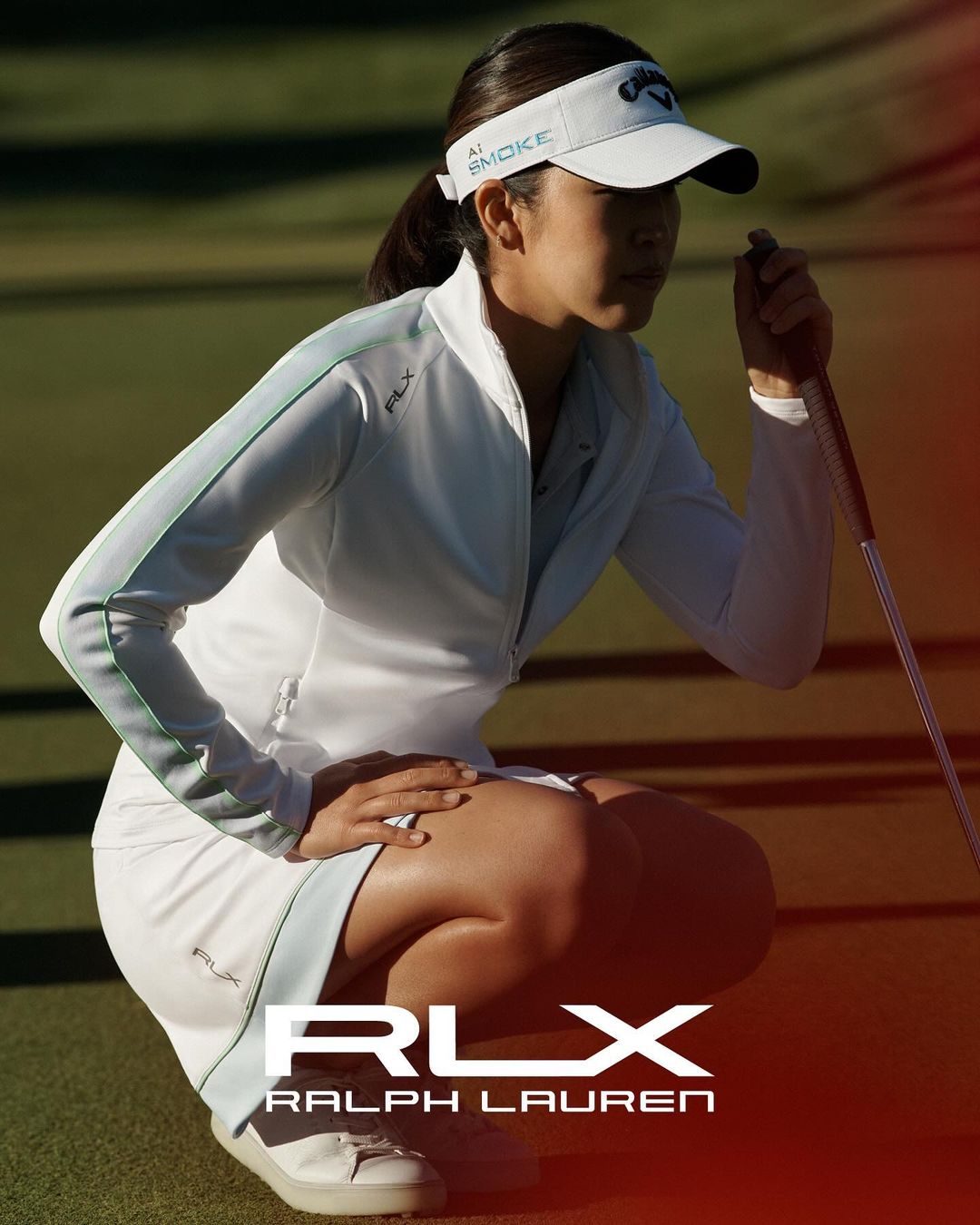 An Asian female golfer in white sportswear, crouching on the green and holding a golf club, outdoors, with Ralph Lauren logo.