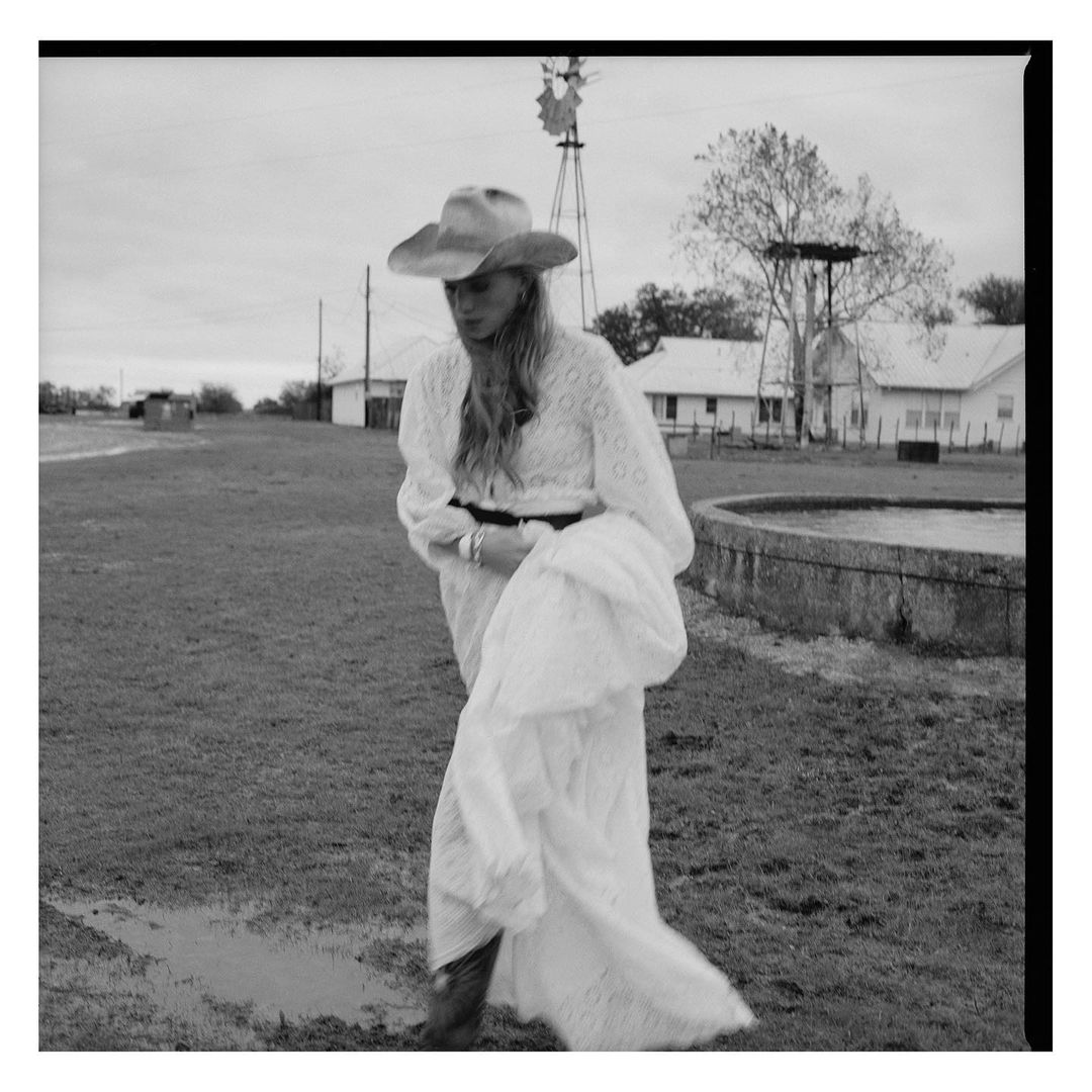 Black and white portrait of a woman in a cowboy hat and white lace dress standing in a grassy field with a windmill in the background.