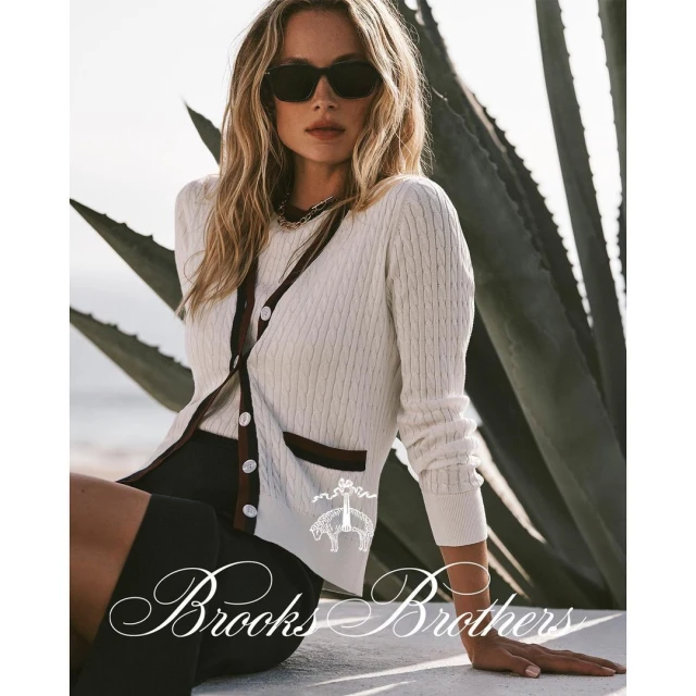 A woman models Brooks Brothers clothing while sitting outside in natural light, wearing sunglasses, a cardigan, and shorts.