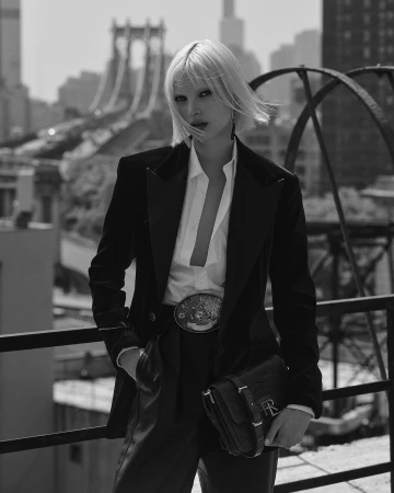 A blonde fashion model poses in a black suit with a white shirt and black leather bag on a rooftop with an urban background in black and white.