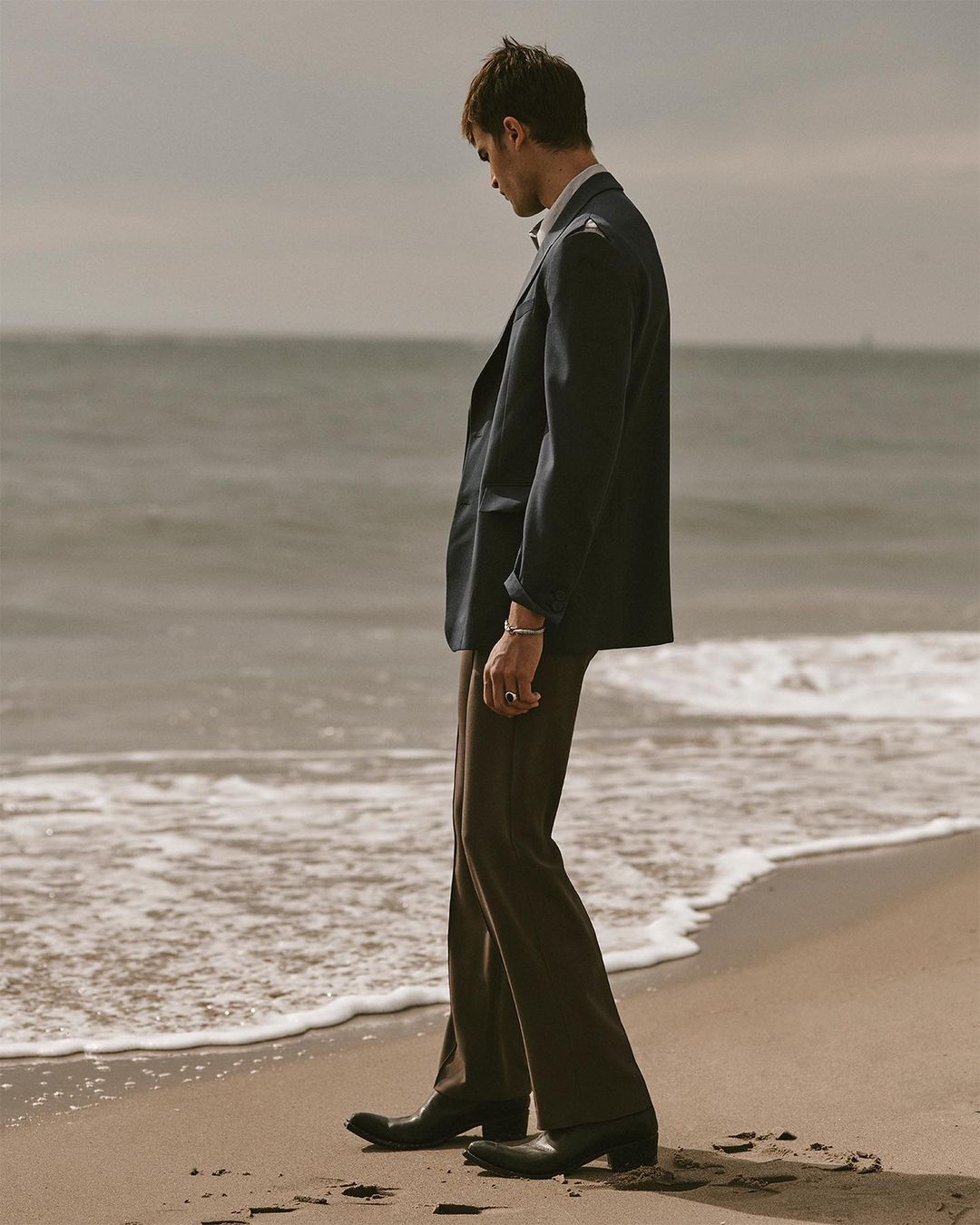 Man in suit jacket and dress pants walks on beach with ocean waves in background, neutral tones, overcast day, fashion editorial style.