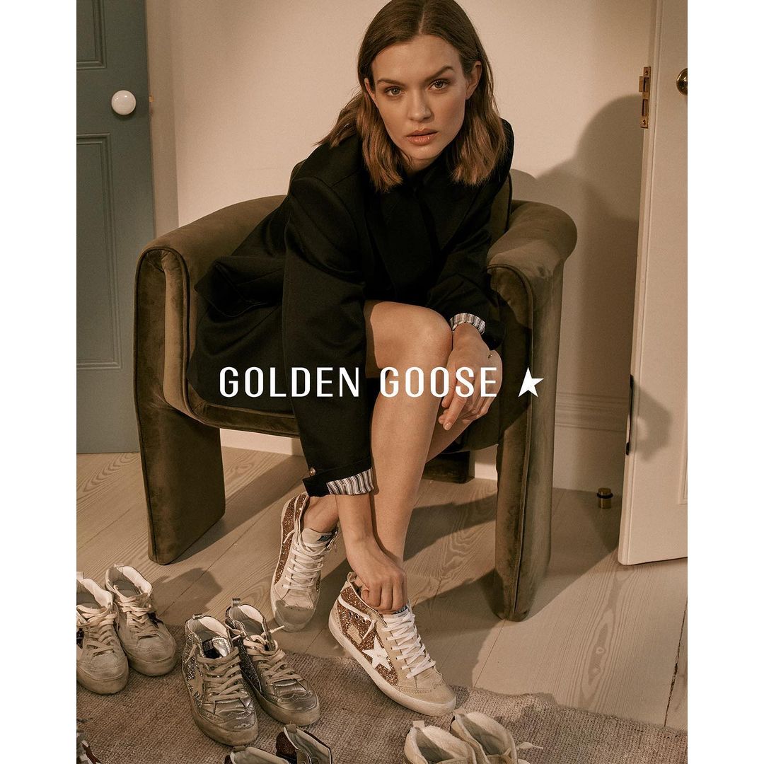 A woman in a black coat sits in a velvet chair, showcasing Golden Goose sneakers. Various pairs of shoes are arranged on the floor.