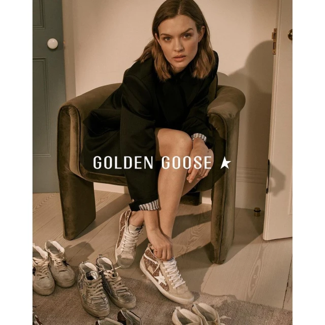 A woman in a black coat sits in a velvet chair, showcasing Golden Goose sneakers. Various pairs of shoes are arranged on the floor.