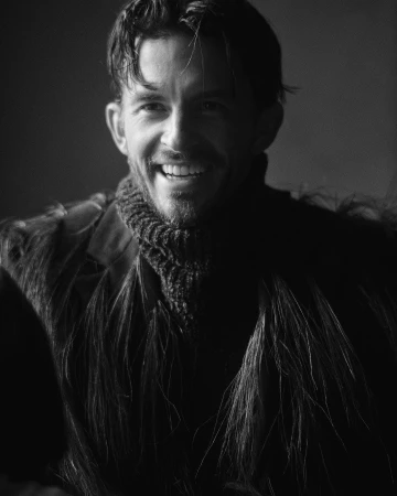 Monochrome portrait of a smiling man wearing a textured turtleneck sweater and dark fringed outerwear.