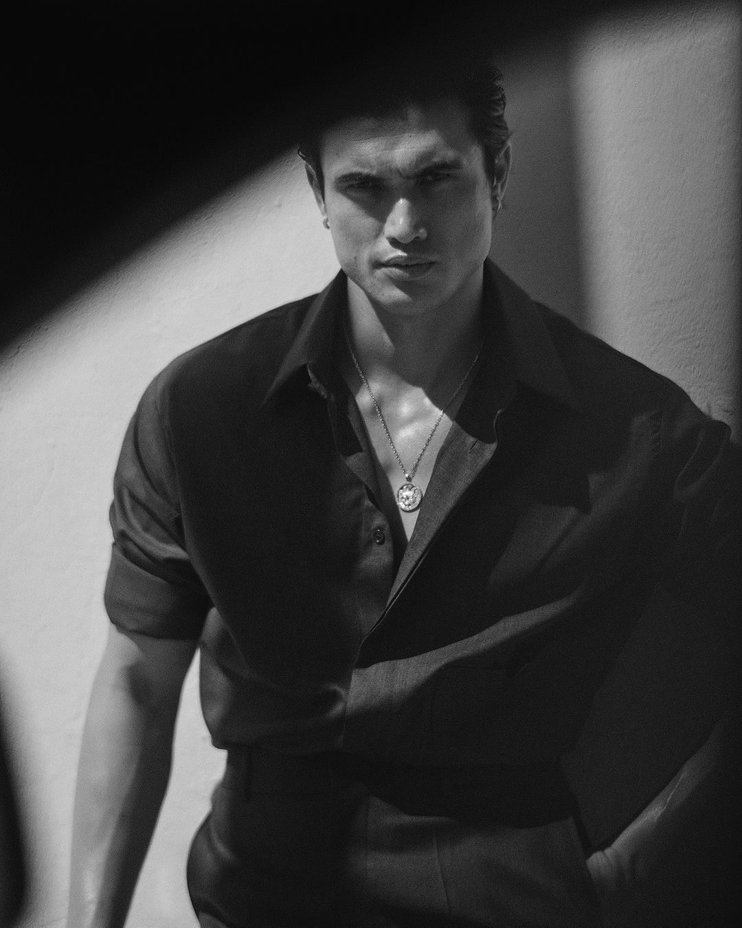 Monochrome portrait of a man in a dark button-down shirt wearing a pendant necklace against a light wall.