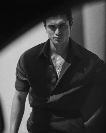 Monochrome portrait of a man in a dark button-down shirt wearing a pendant necklace against a light wall.