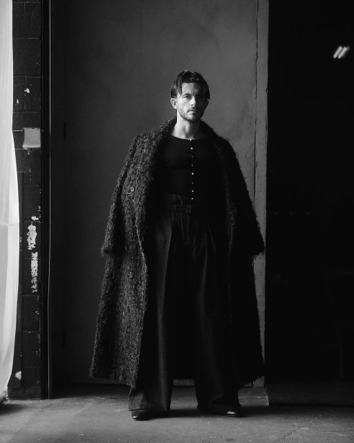 Black and white portrait of a man wearing a dark shirt and long coat, standing in front of a textured wall and doorway in dim lighting.