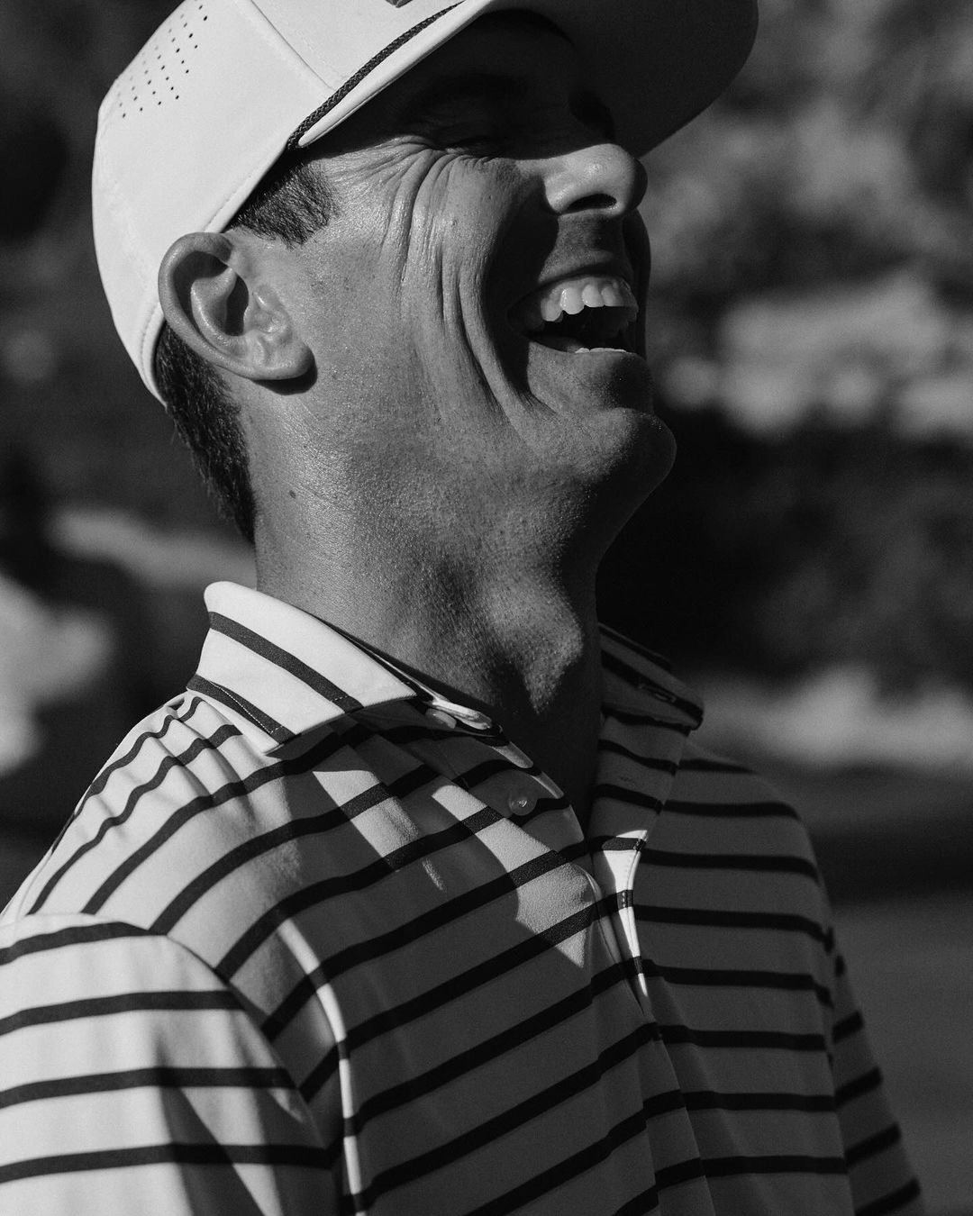 Monochrome portrait of a smiling man in a striped polo shirt and baseball cap, side view, in natural outdoor light with blurred background.