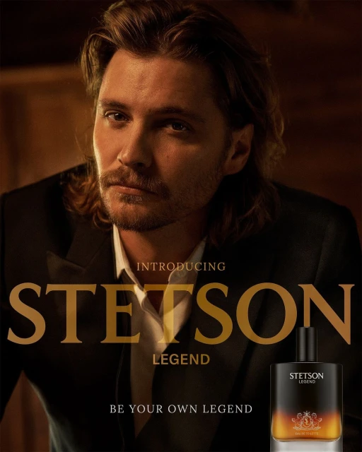 A man with long hair poses in a black suit next to Stetson Legend cologne. The image is dark and moody with warm, golden light.