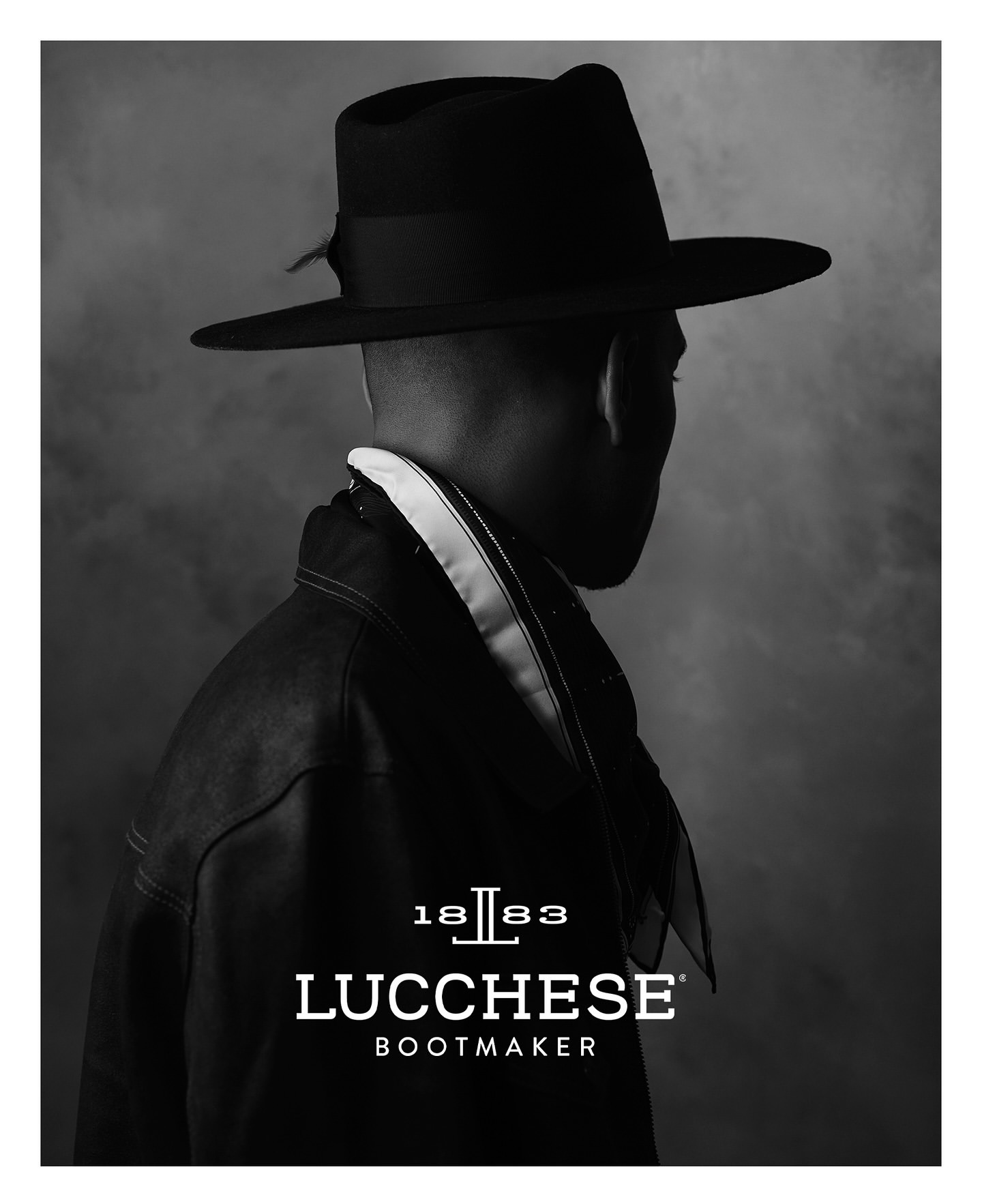 Monochrome portrait of a person wearing a fedora and leather jacket, featuring the Lucchese Bootmaker logo against a blurred background.