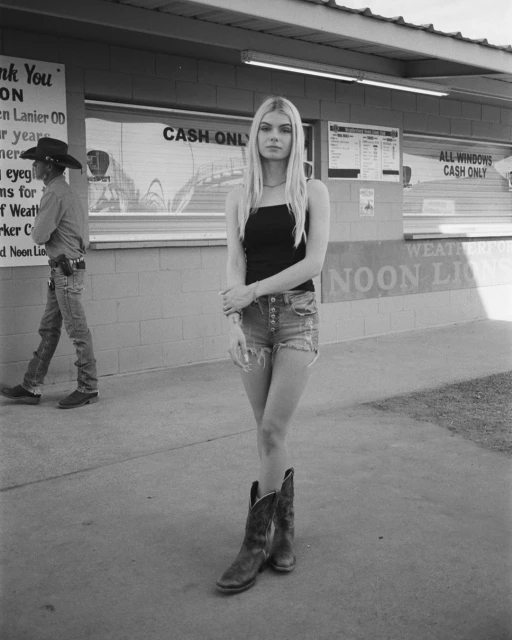 Black and white portrait of a young woman in denim shorts and cowboy boots, standing outside a rural building with a cowboy walking by.