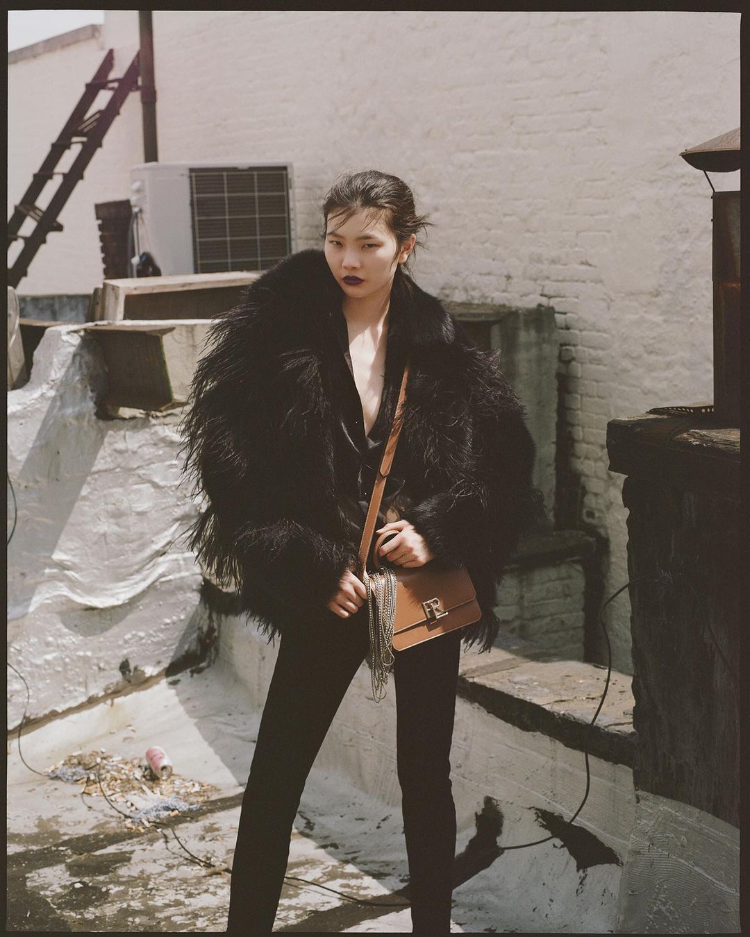 Fashion shot of a young Asian woman wearing a black fur coat, black pants, and a brown leather purse in an urban setting.