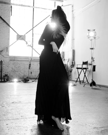Black and white portrait of a model posing in a long dark dress in a studio with backlighting and studio equipment