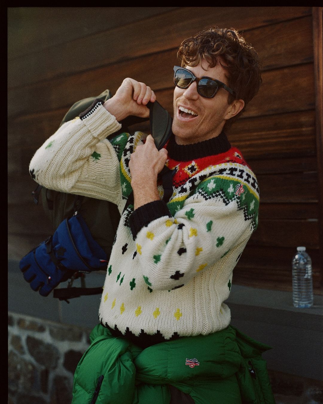 Smiling adult man wearing a patterned sweater and sunglasses, zipping up a backpack, standing outside on a stone stoop near a wooden wall.