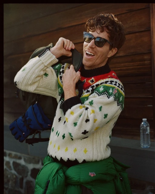 Smiling adult man wearing a patterned sweater and sunglasses, zipping up a backpack, standing outside on a stone stoop near a wooden wall.