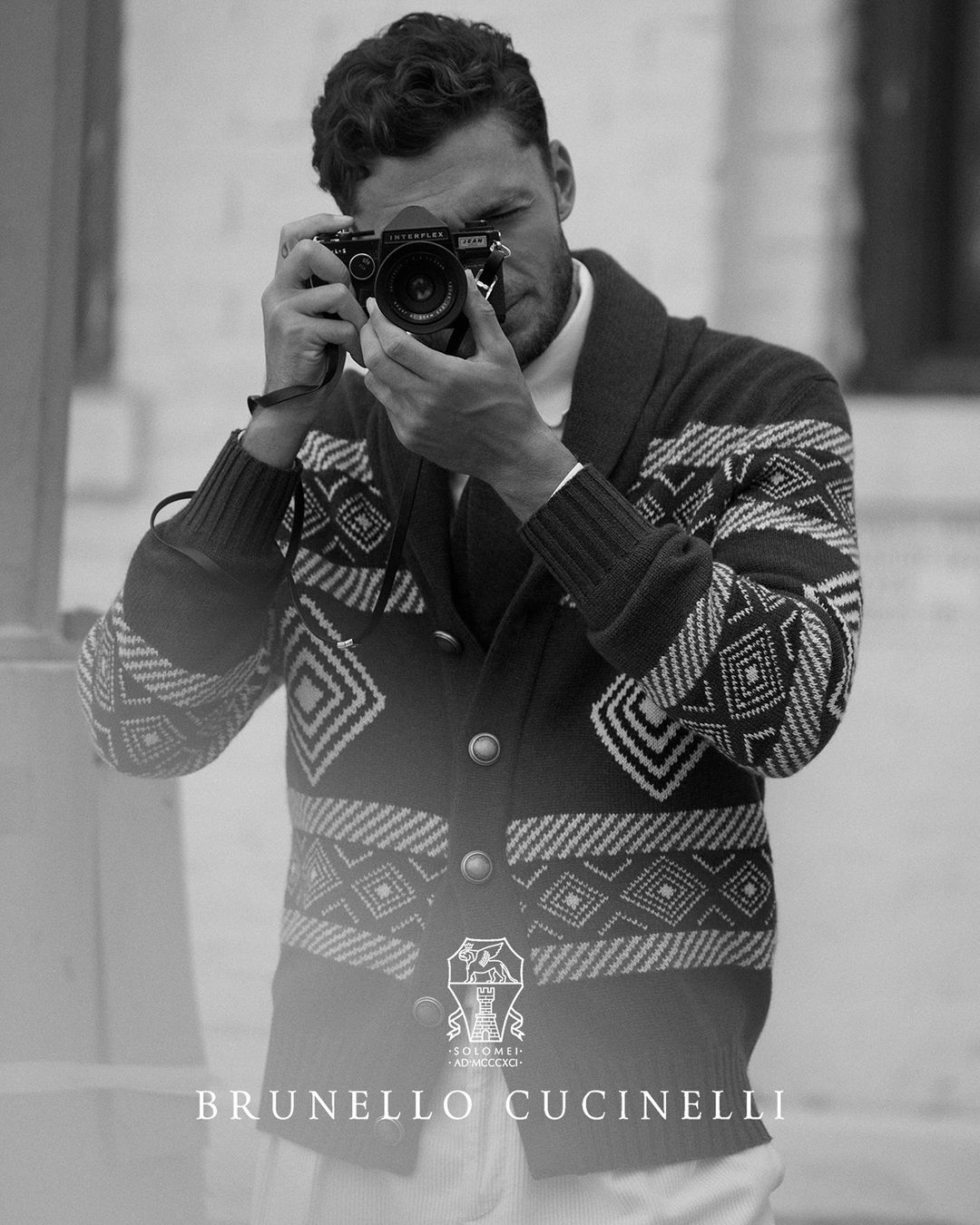 Monochrome portrait of a man with a digital camera, wearing a patterned cardigan; Brunello Cucinelli logo visible at the bottom.