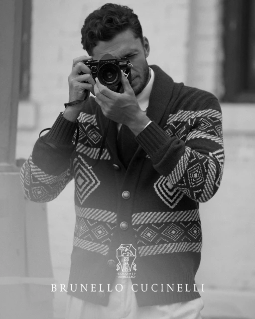 Monochrome portrait of a man with a digital camera, wearing a patterned cardigan; Brunello Cucinelli logo visible at the bottom.