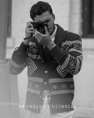 Monochrome portrait of a man with a digital camera, wearing a patterned cardigan; Brunello Cucinelli logo visible at the bottom.