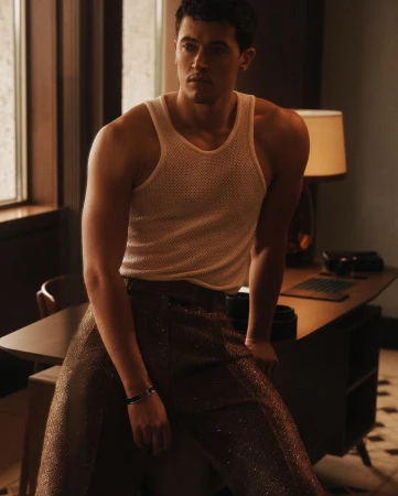 Man wearing a cream knit tank top and dark metallic trousers leaning against a desk in a moody room by Blair Getz Mezibov