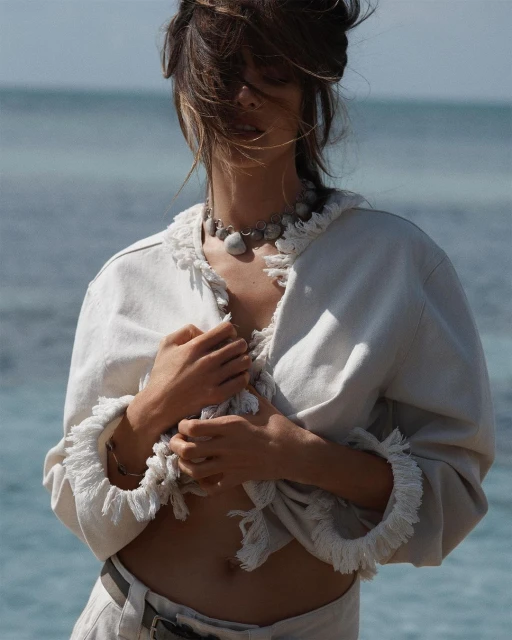 A woman on a beach wears a white jacket with fringe detail, beige pants, and a shell necklace; the ocean is in the background.