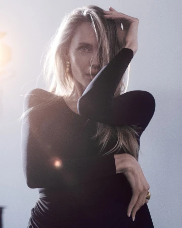 Portrait of a blonde woman wearing a dark long-sleeve top, posing with her hand in her hair against a neutral light background.