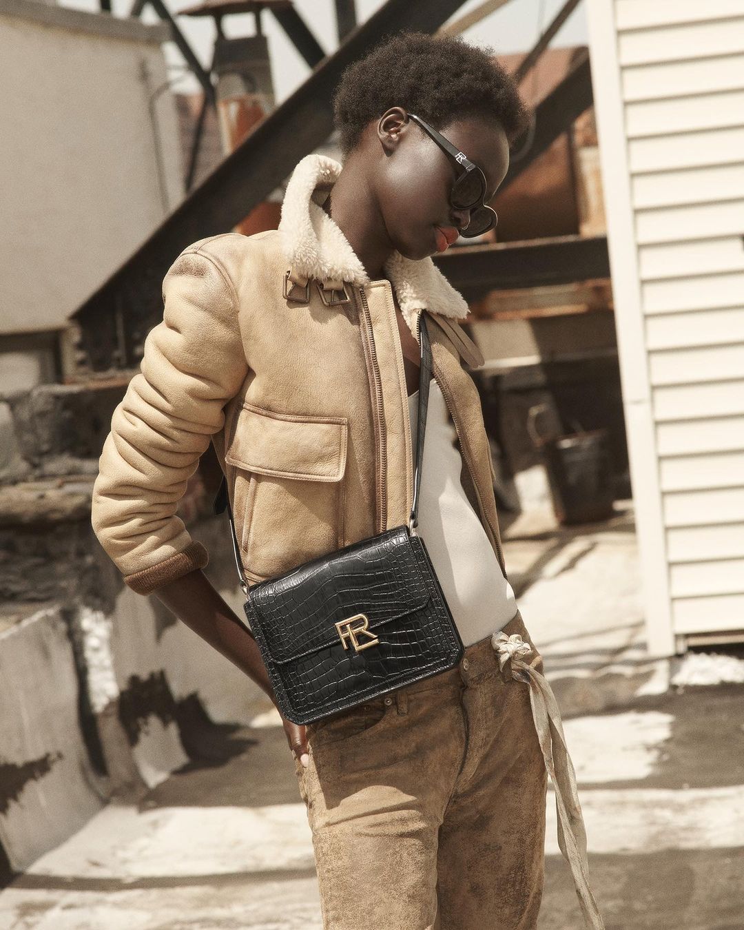 A woman wearing sunglasses, a shearling jacket, suede pants, and a leather purse with a gold buckle poses in front of a white building.