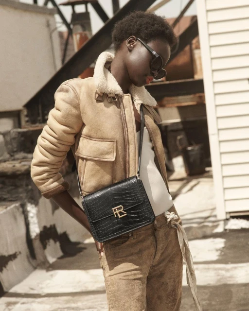 A woman wearing sunglasses, a shearling jacket, suede pants, and a leather purse with a gold buckle poses in front of a white building.