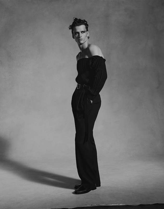Monochrome fashion shot of a slender adult male model with dark eye makeup in an off-the-shoulder top and trousers against a grey backdrop.