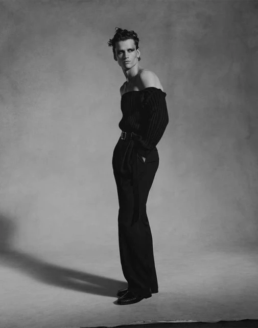 Monochrome fashion shot of a slender adult male model with dark eye makeup in an off-the-shoulder top and trousers against a grey backdrop.