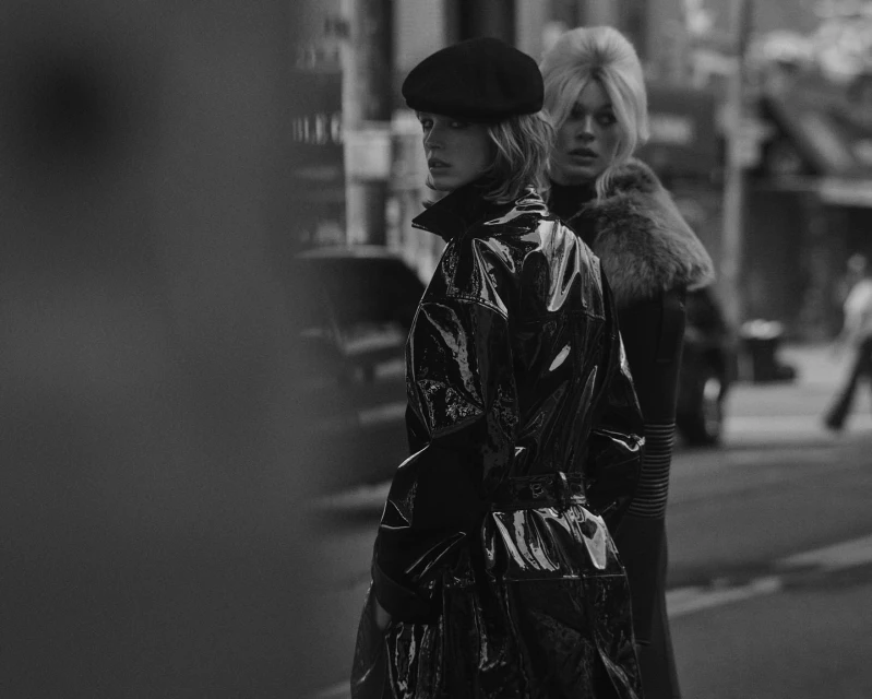 Two women in black fashionable coats and hats stand on a city street. The woman in the front wears a shiny coat, and the other has a fur collar.