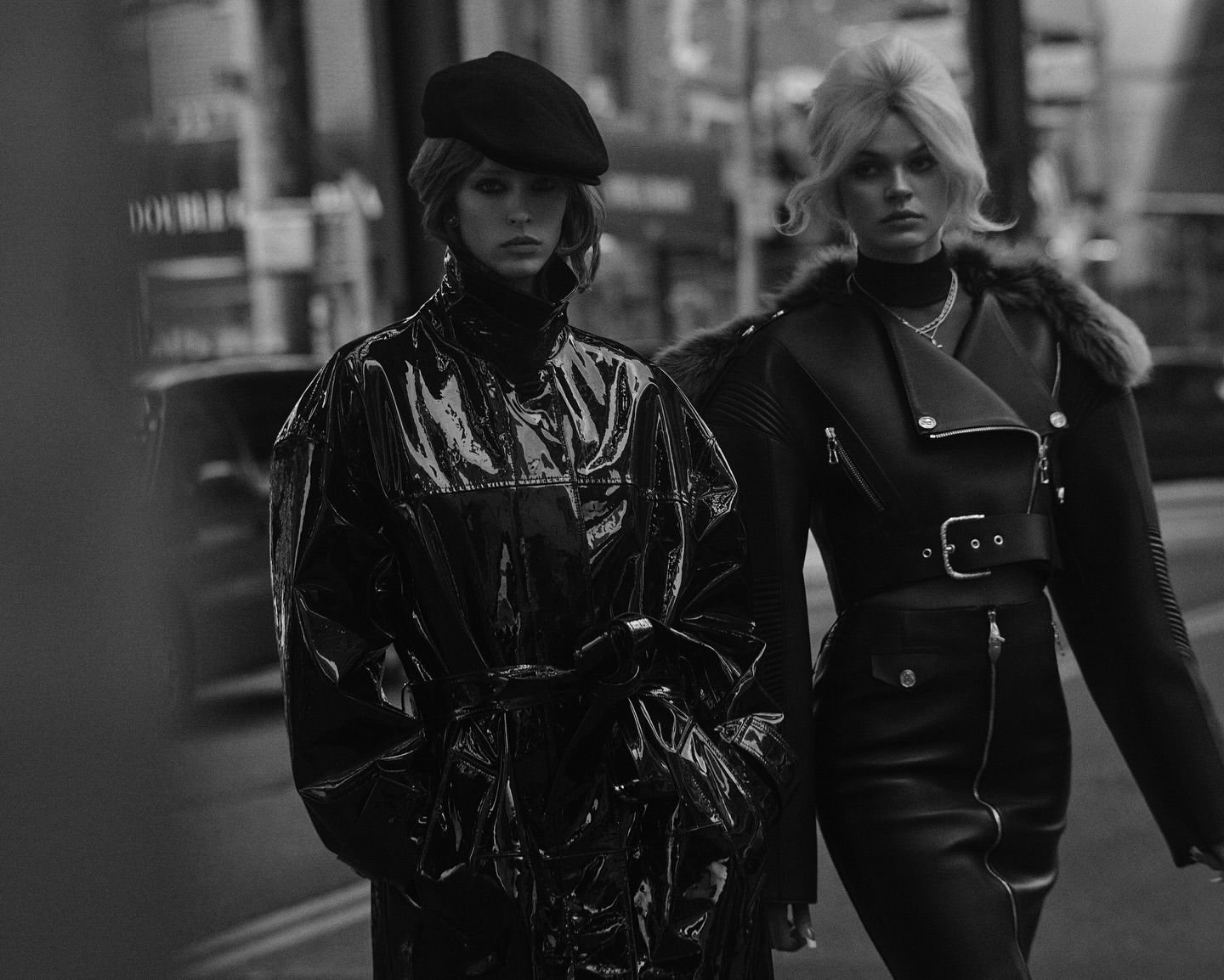 Two women in black coats on city street, monochrome image featuring dark fashion and urban setting, captured in a medium shot.