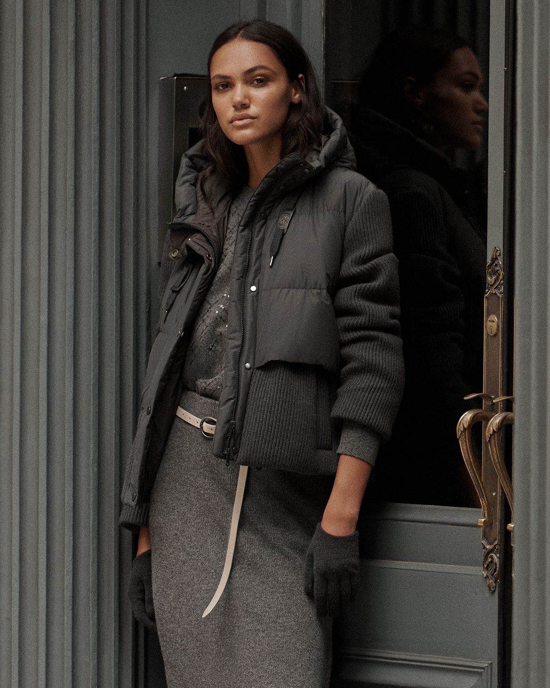 A woman stands in front of a gray door wearing a gray dress, puffer jacket, and matching gloves in a medium, studio shot.