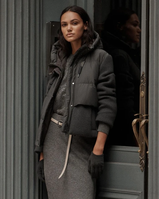 A woman stands in front of a gray door wearing a gray dress, puffer jacket, and matching gloves in a medium, studio shot.