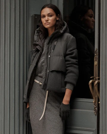 A woman stands in front of a gray door wearing a gray dress, puffer jacket, and matching gloves in a medium, studio shot.