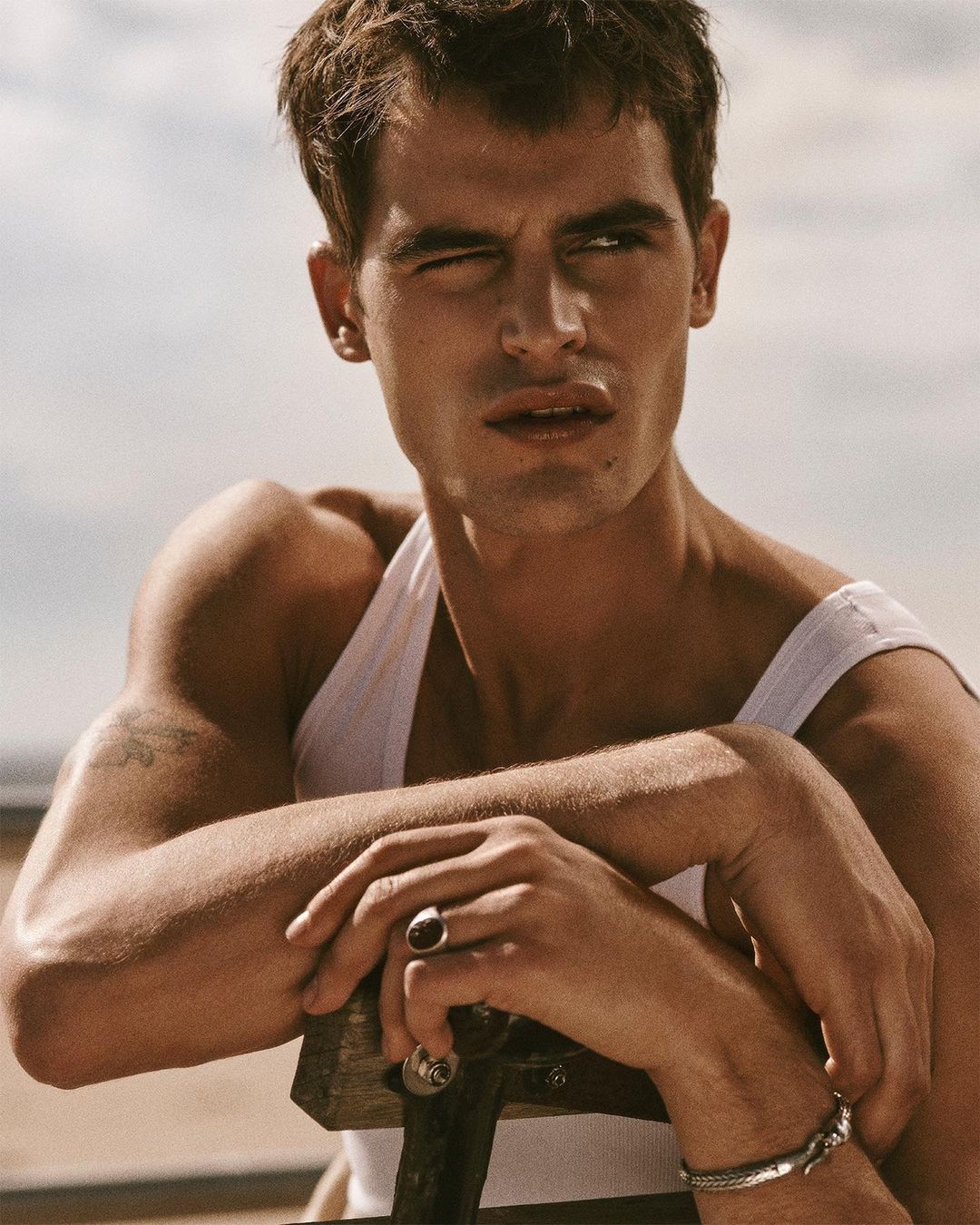 Portrait of adult male with brown hair wearing a white tank top, silver jewelry, and a ring, against a neutral outdoor background.