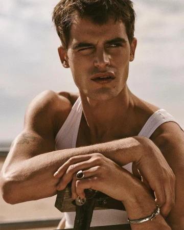 Portrait of adult male with brown hair wearing a white tank top, silver jewelry, and a ring, against a neutral outdoor background.