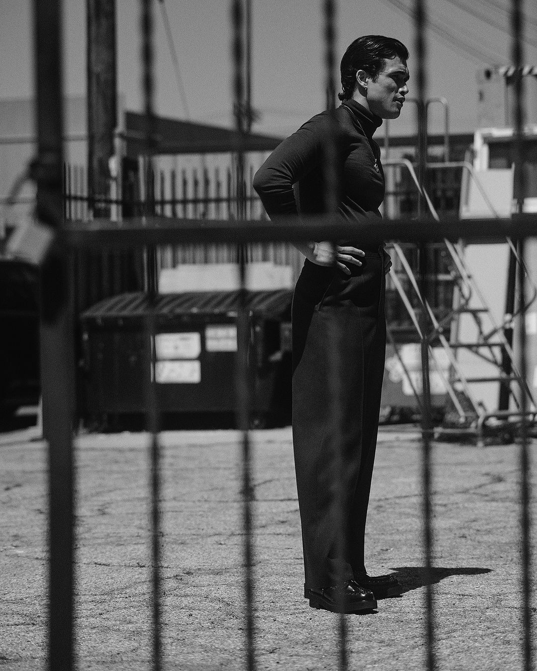 Monochrome image of a man posing outdoors in a dark turtleneck and pants with a metal fence, ladder, and dumpster in the background.