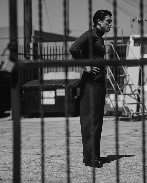 Monochrome image of a man posing outdoors in a dark turtleneck and pants with a metal fence, ladder, and dumpster in the background.