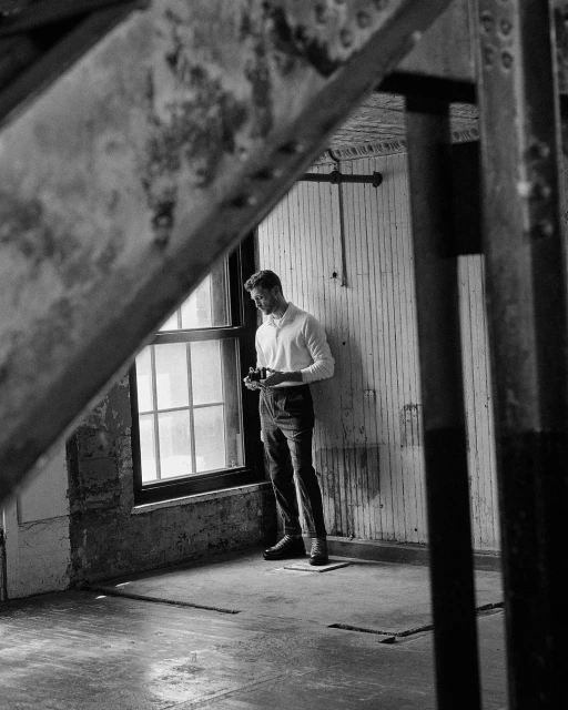 Black and white shot of a man in an empty industrial interior, holding a vintage camera near a window with riveted metal beams.