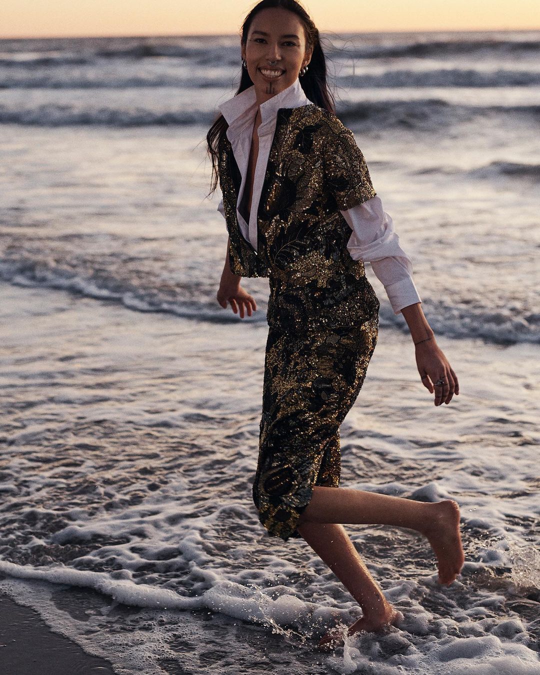 A woman in a gold sequin outfit is walking barefoot through the water on a beach at sunset with a serene and joyful expression.