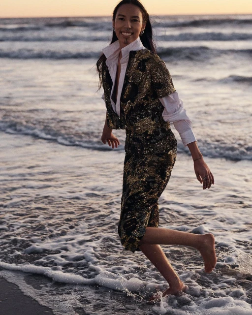 A woman in a gold sequin outfit is walking barefoot through the water on a beach at sunset with a serene and joyful expression.
