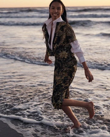 A woman in a gold sequin outfit is walking barefoot through the water on a beach at sunset with a serene and joyful expression.