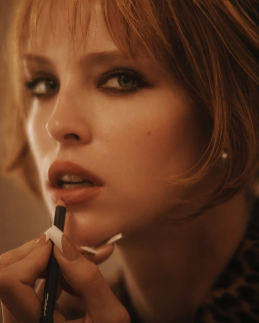 Close-up of a young adult female model applying lip liner with a makeup pencil in a warm, dimly lit beauty or fashion setting.