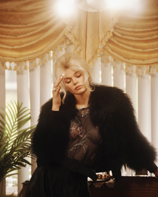 Woman with blonde hair wearing fur coat resting hand on head indoors near window with gold curtains in a medium, eye-level studio shot.