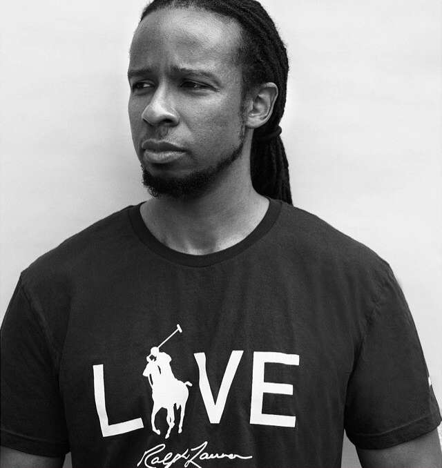 Portrait of an adult African American man in a black Ralph Lauren t-shirt with dreadlocks, looking off to the side against a plain background.
