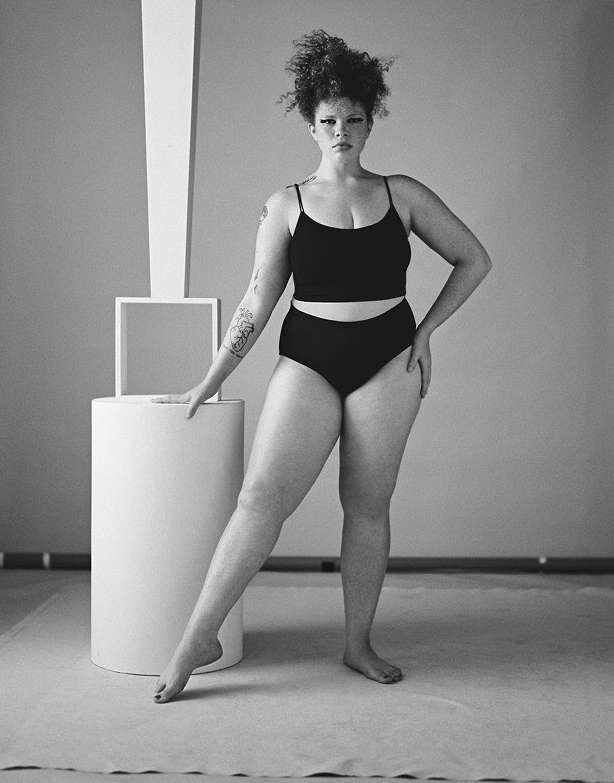 Monochrome studio shot of a plus-size adult woman in dark two-piece swimwear, posing near a white geometric prop.